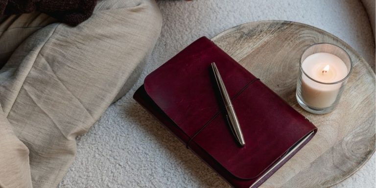 Journal de cuero Muse burdeos sobre bandeja de madera, con bolígrafo encima y vela encendida.