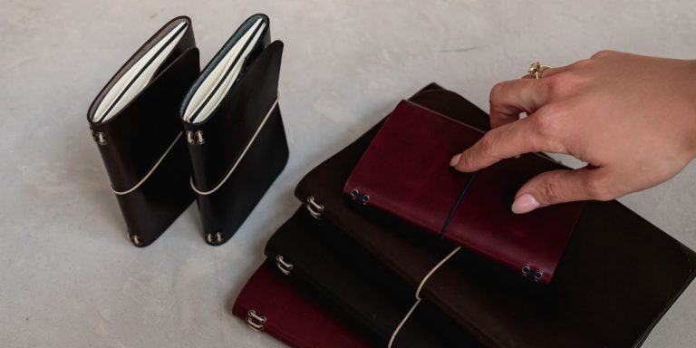 Dos journals de cuero en vertical y una pila de journals en burdeos y marrón; una mano señala el journal superior sobre un fondo neutro.