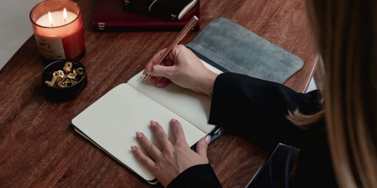 Manos escribiendo en un journal abierto sobre una mesa de madera, con una vela encendida y un cuenco con pinzas metálicas al lado.