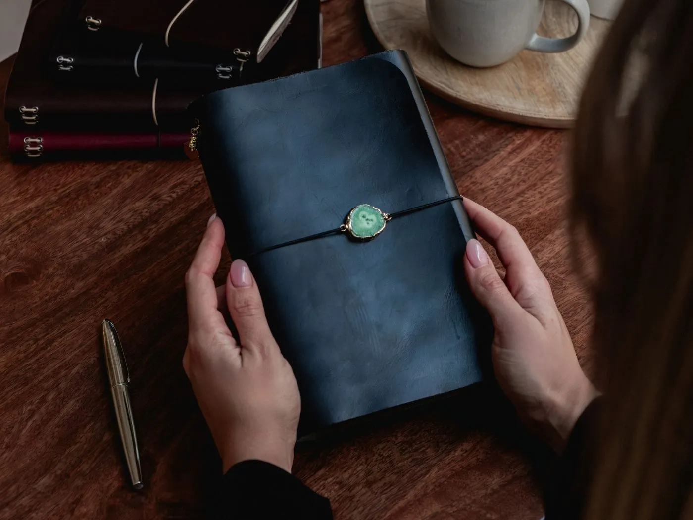 Manos sosteniendo un journal de cuero notāre con charm conector verde sobre una mesa con café