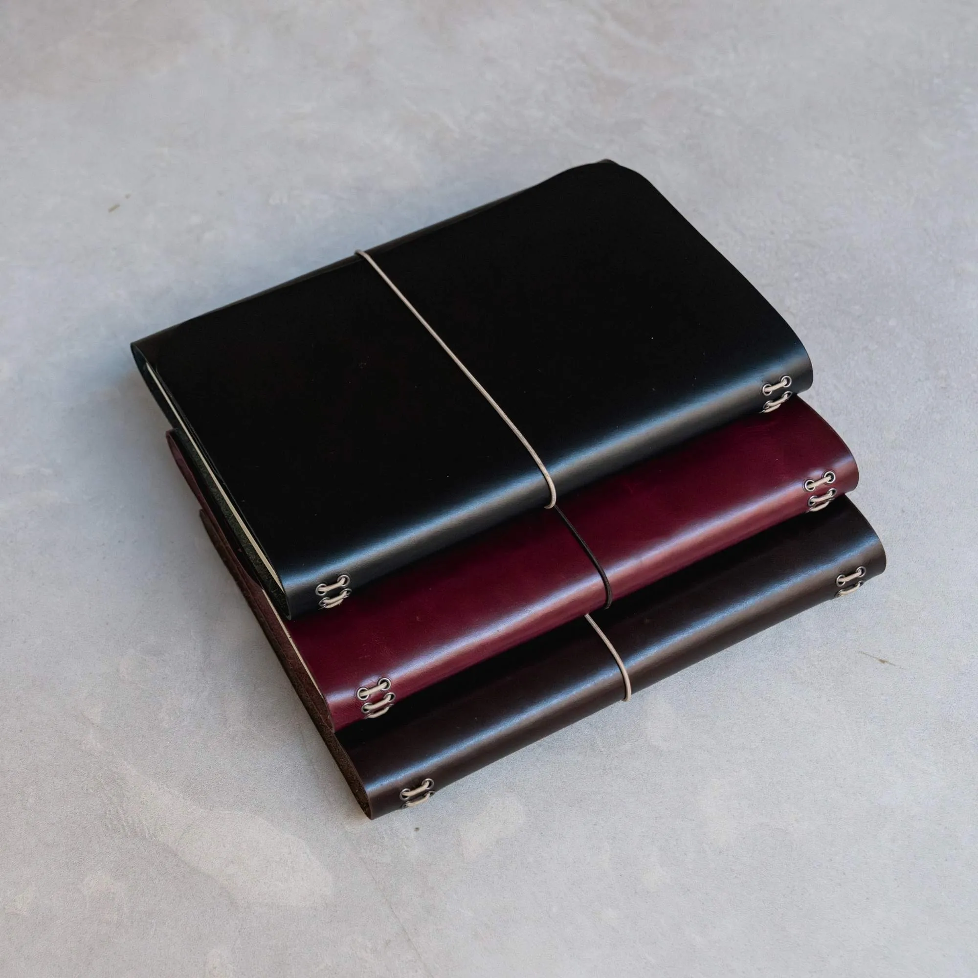Journals notāre de cuero apilados en tres colores (negro, burdeos y marrón), con elásticos y ojales metálicos.