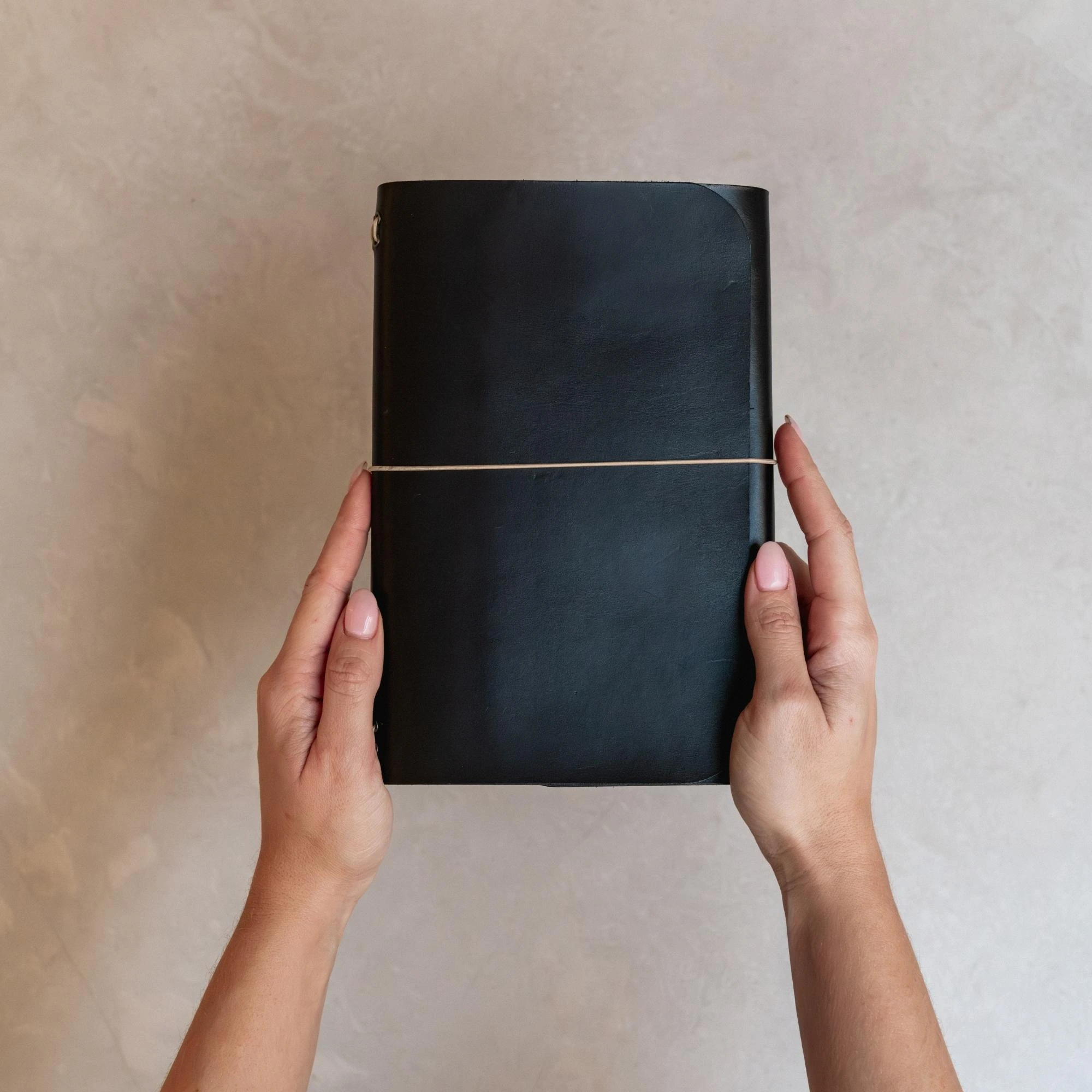 Manos sujetando un journal de cuero negro notāre con cierre elástico, sobre fondo neutro.