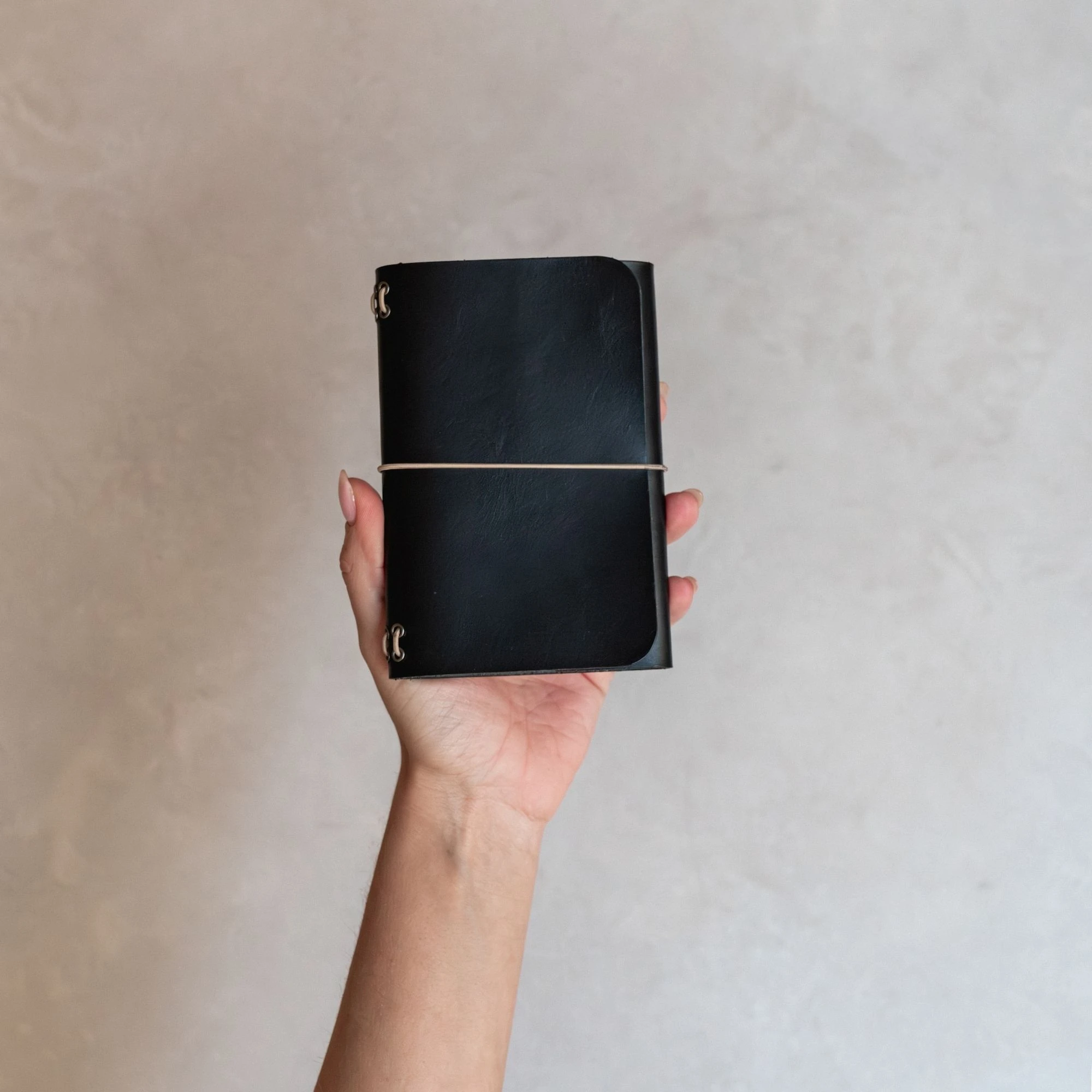 Mano sujetando un journal de cuero negro notāre con cierre elástico, sobre fondo neutro.