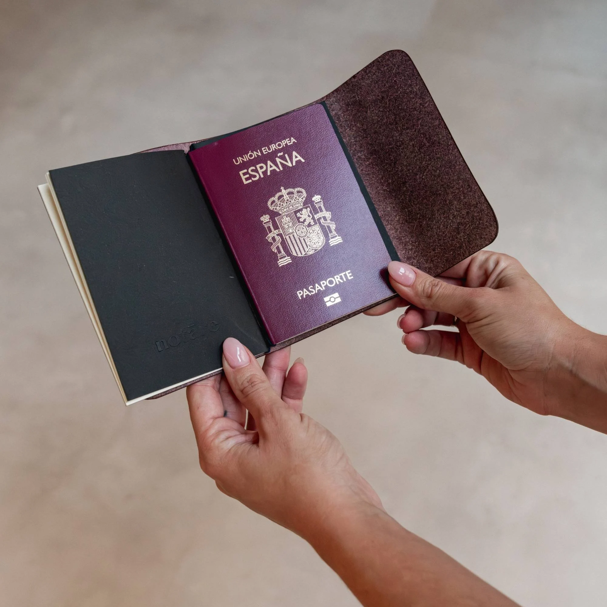 Manos sosteniendo un notāre Nomad abierto con un pasaporte español encajado en el interior, sobre fondo neutro.
